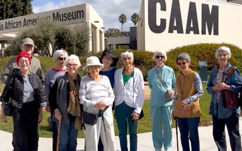 A group of older adults poses and smiles in front of the California African American Museum (CAAM) on a sunny day. Some people wear hats and sunglasses, and a few hold canes.