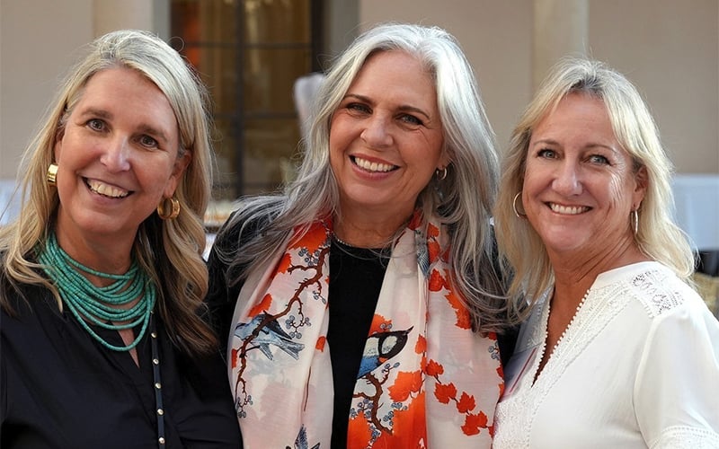 Three smiling women pose together outdoors. The woman in the middle has long gray hair and wears a floral scarf, while the women on either side have blonde hair and wear necklaces and light-colored tops.