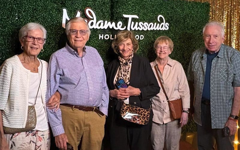 Five older adults stand smiling together in front of a green backdrop with the “Madame Tussauds Hollywood” logo. Four are holding handbags, and they are dressed in casual, comfortable clothing.
