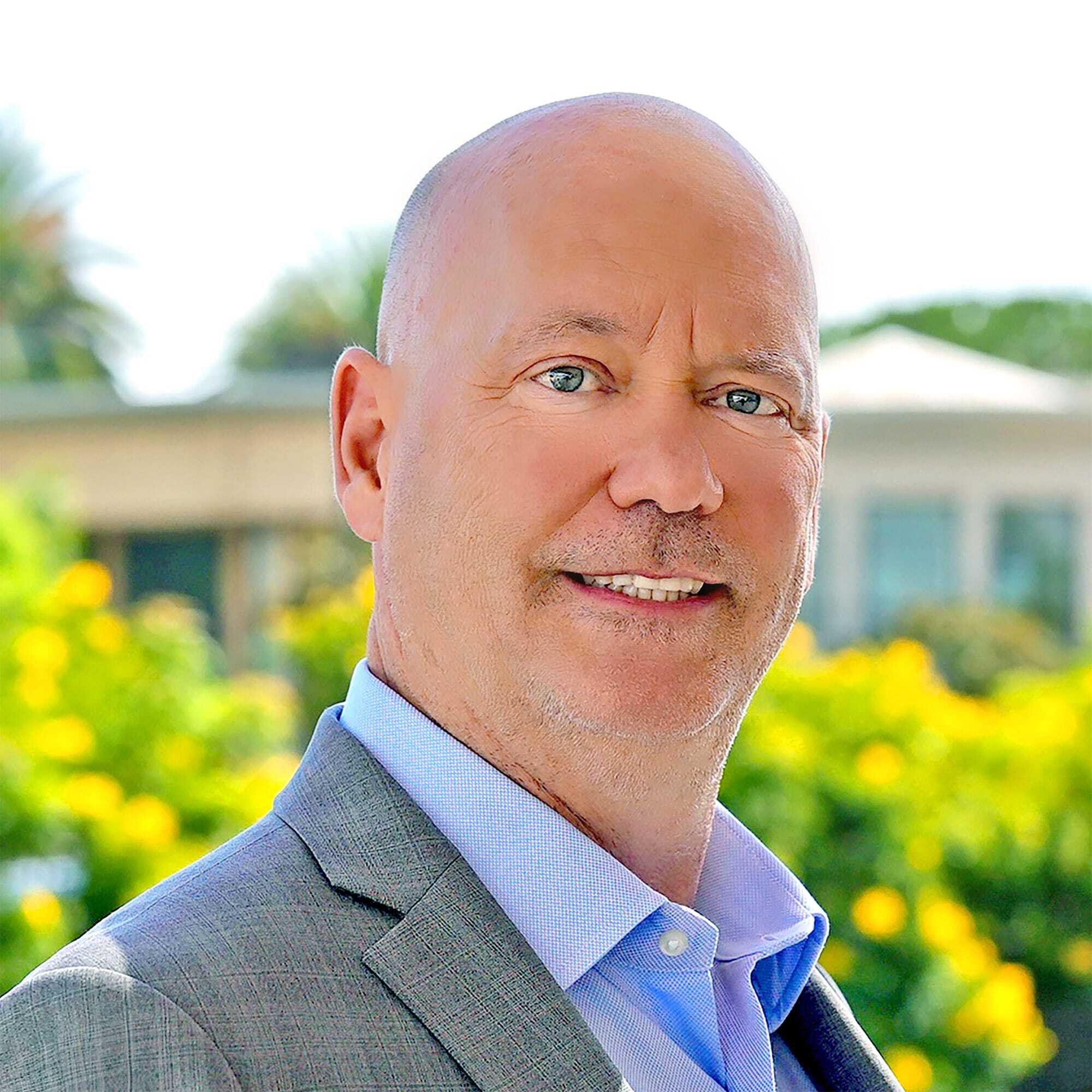 A middle-aged man with a shaved head wearing a grey suit jacket and light blue shirt, smiling outdoors with greenery and a building blurred in the background.