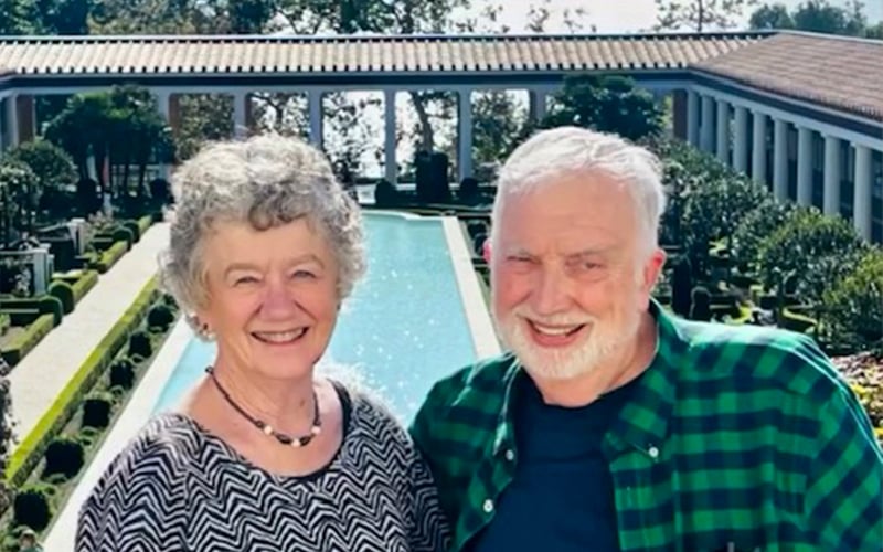 An older smiling couple poses in front of a long rectangular reflecting pool, surrounded by manicured gardens and colonnades, on a sunny day.