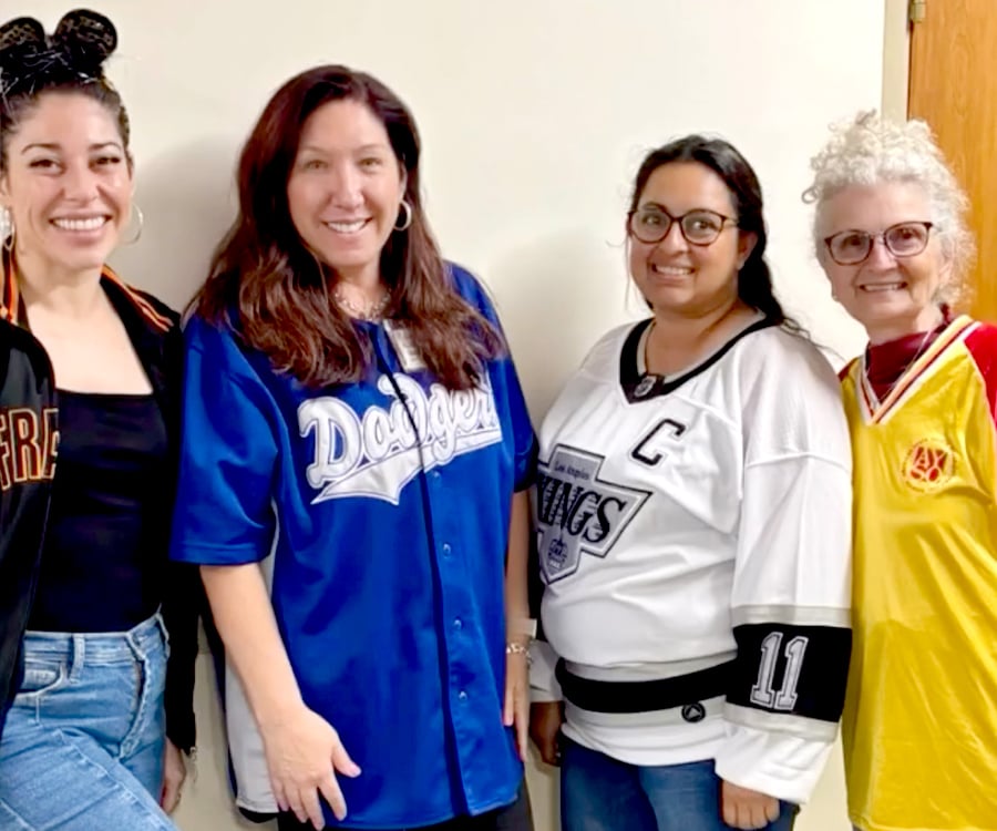 Four women stand together smiling, each wearing a different sports jersey, including teams in blue, black, white, and yellow, against a plain indoor background.