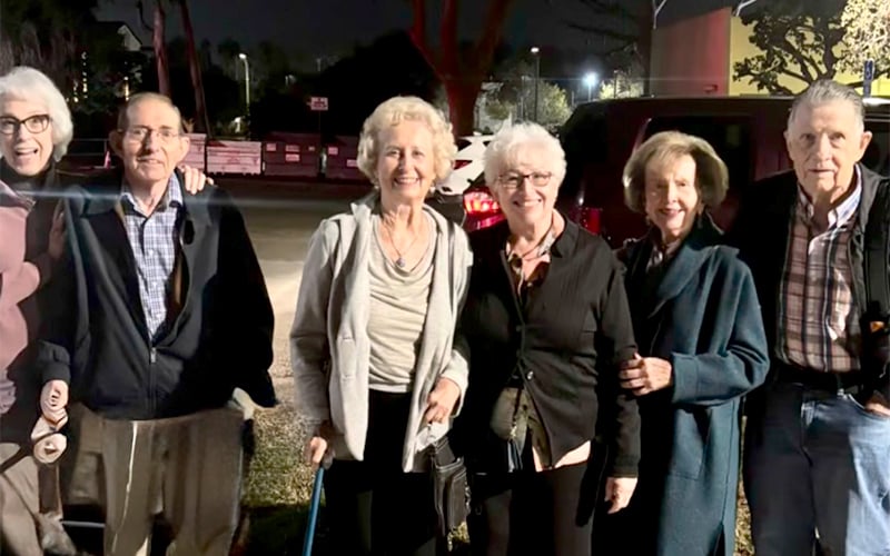 Six older adults stand together smiling outdoors at night, dressed warmly in jackets and sweaters. A car and trees are visible in the background under streetlights.