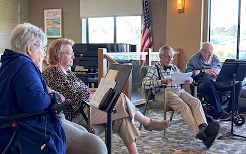 Four older adults sit in a circle indoors, reading from papers or booklets. An American flag, piano, and large window are in the background. The group appears to be engaged in a discussion or activity.