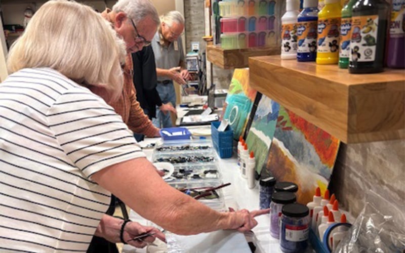 Several adults work at a table filled with art supplies, including paint, brushes, and bottles of glue, as they engage in a painting activity. Colorful paintings and art materials are visible on shelves and the table.