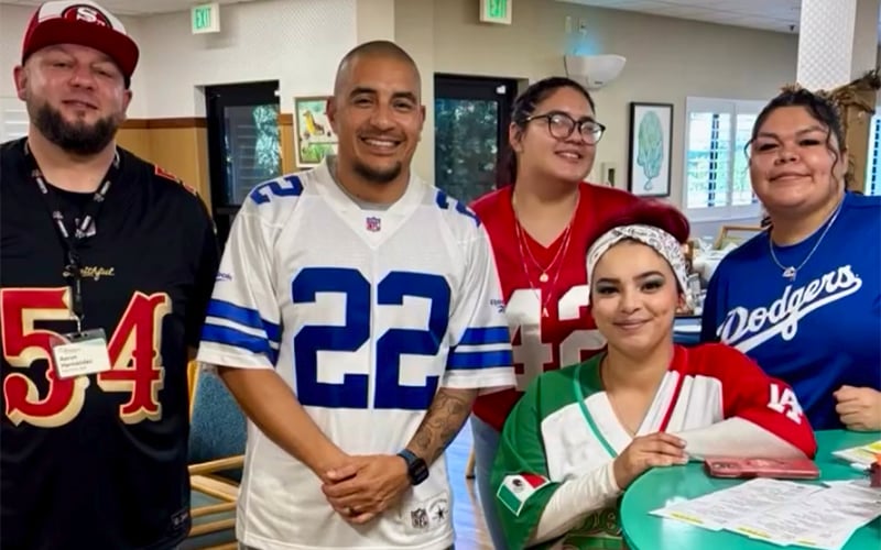 Five smiling people pose indoors wearing sports jerseys, including the 49ers, Cowboys, and Dodgers. One person holds papers at a table. The background shows chairs and green exit signs.