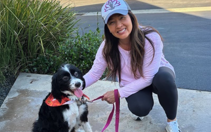 A woman in a light purple hat and shirt kneels on a sidewalk, smiling beside a black and white dog wearing an orange bandana and pink leash. Green bushes and pavement are in the background.