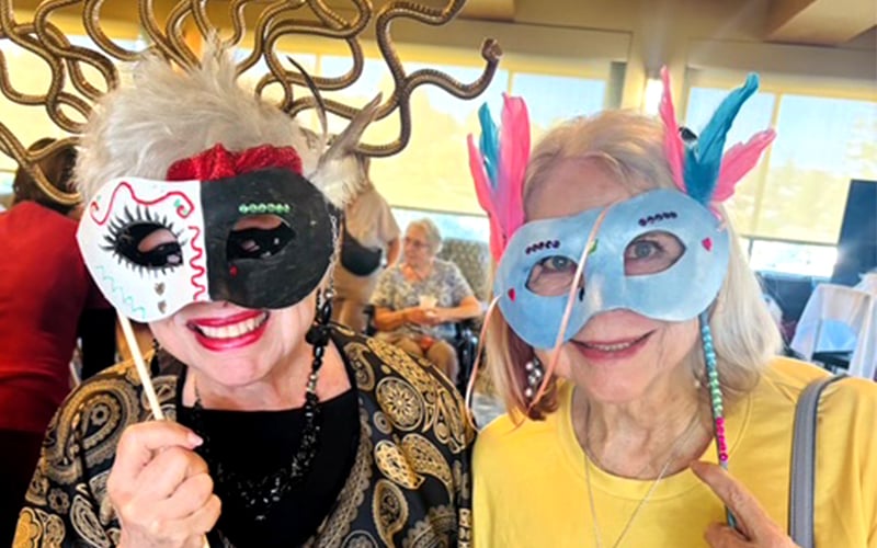 Two smiling older women pose wearing colorful masquerade masks; one has a costume with gold headpiece shaped like snakes, the other wears a mask with attached feathers. They appear to be at a festive indoor event.
