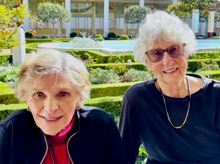Two elderly women with white hair smile at the camera while sitting outside in a sunny, neatly landscaped garden with trimmed hedges, green plants, and a fountain in the background.