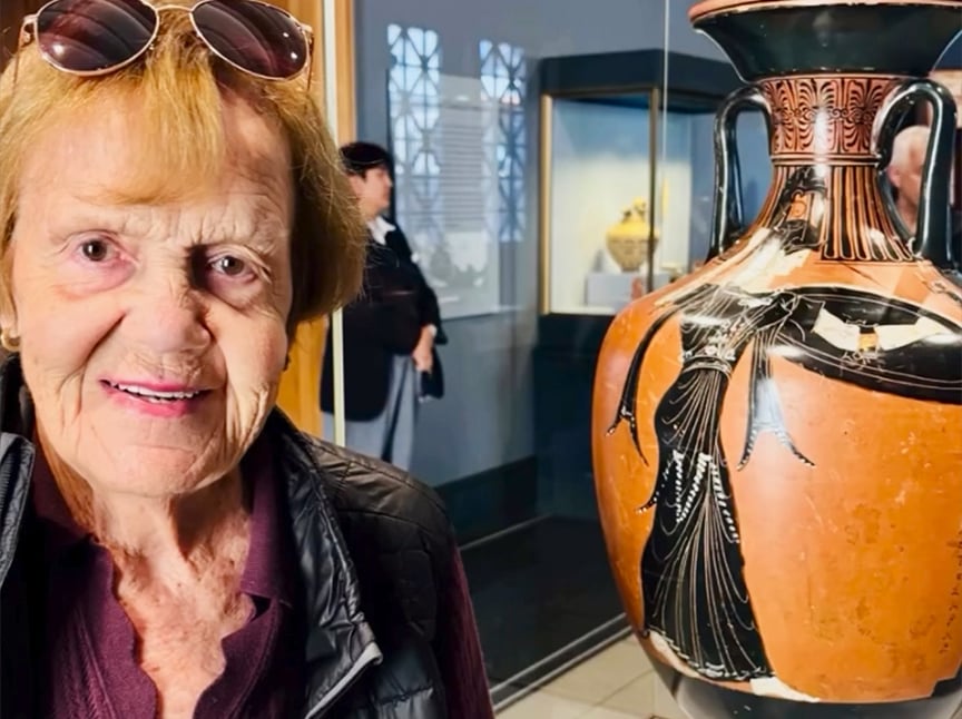 An older woman smiles beside a large ancient Greek vase with black-figure artwork, displayed in a museum setting with other visitors and artifacts visible in the background.