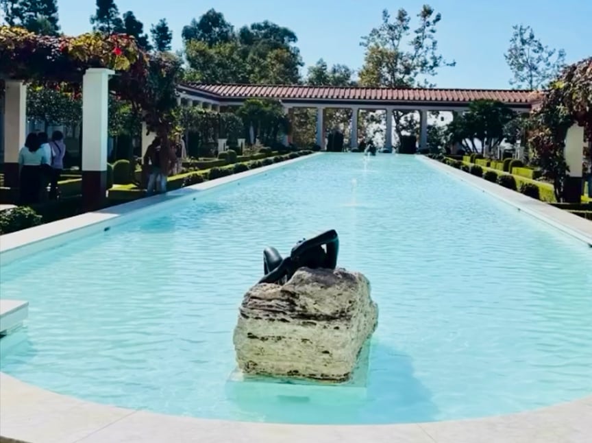 A long rectangular reflecting pool with a sculpture on a rock in the center, surrounded by columns, gardens, and hedges under a clear blue sky. People are walking along the pool’s edge.