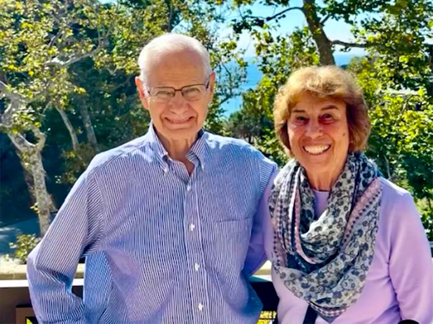 An older man and woman stand smiling together outdoors in front of leafy trees. The man wears glasses and a striped shirt, and the woman wears a patterned scarf and sweater. Sunlight brightens the scene.