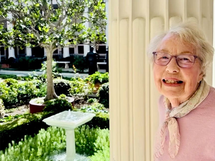 An elderly woman with glasses and a scarf smiles beside a column, with a lush garden, a tree, and a small fountain in the background. Sunlight brightens the peaceful outdoor courtyard scene.