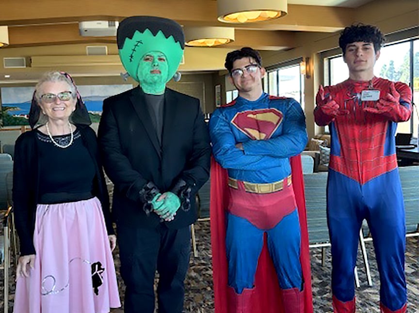 Four people in costumes stand indoors: an older woman in a poodle skirt, a person dressed as Frankensteins monster, one as Superman, and one as Spider-Man. They are posing and smiling for the camera in a bright room.