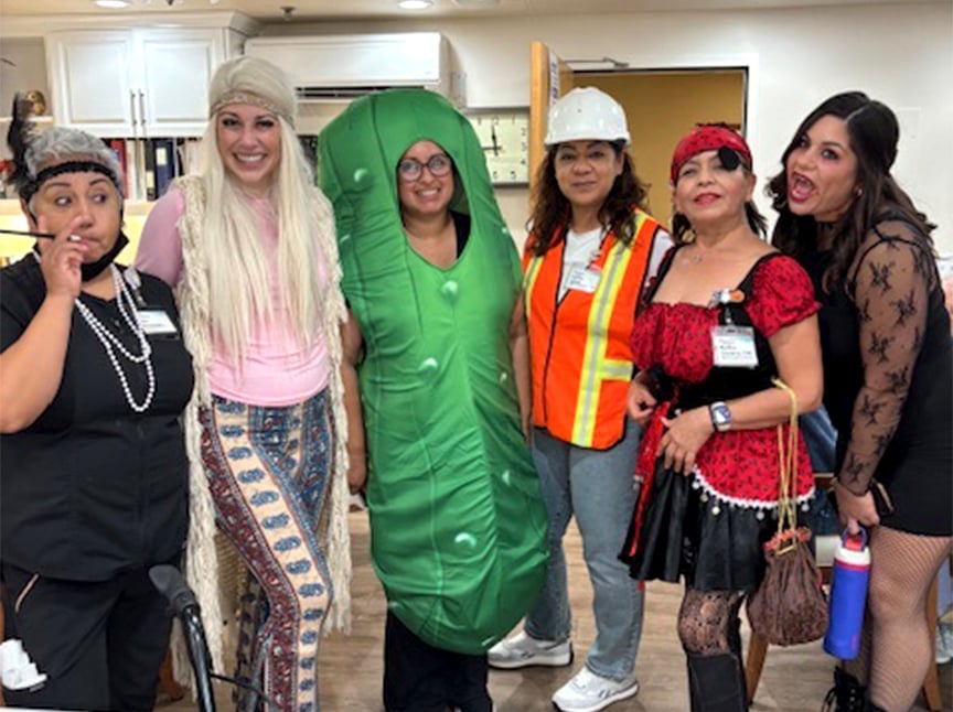 Six women stand together indoors wearing various costumes, including a pickle suit, construction worker, flapper, hippie, flamenco dancer, and a black lace dress. They are smiling and posing for the photo.
