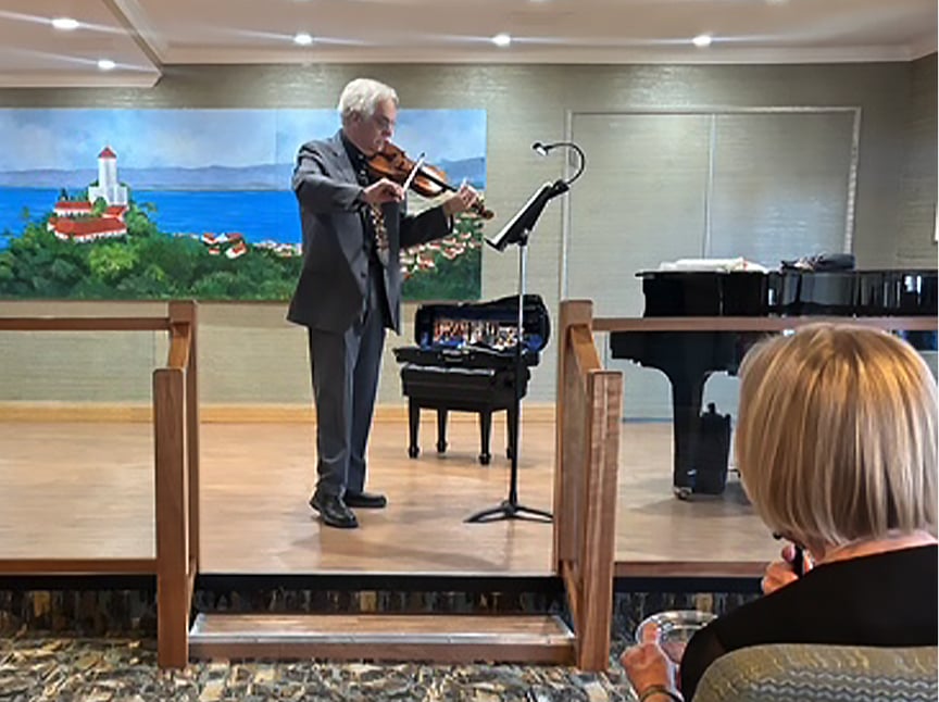 A man in a suit plays the violin on a small indoor stage next to a grand piano, with an audience member watching. A mural of a coastal scene with a lighthouse is on the wall behind him.