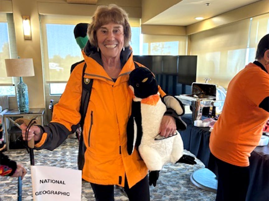 A smiling woman in an orange jacket holds a stuffed penguin and a National Geographic bag while standing indoors at an event. Other people and food tables are visible in the background.