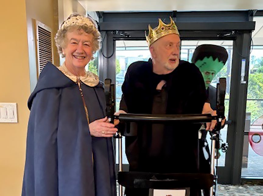 An older woman in a blue cape and bonnet stands smiling next to an older man using a walker, dressed as a king with a crown and black cape. A person in a Frankenstein costume is visible in the background.