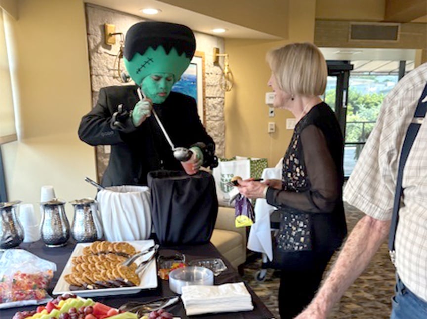 A person dressed as Frankenstein’s monster serves punch to a woman at a buffet table with fruit, cookies, and drinks in a bright indoor setting.