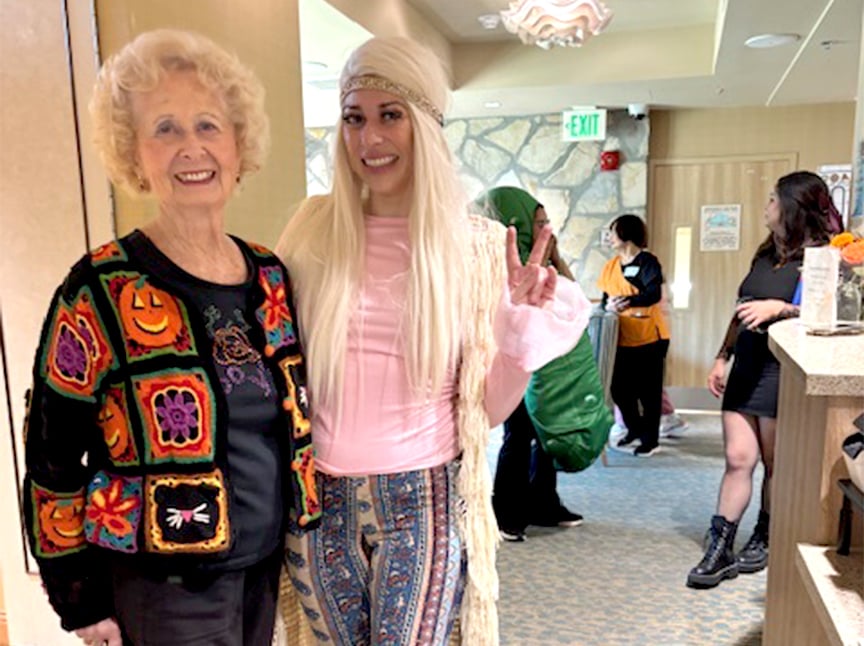 Two women pose indoors; one older woman wears a black sweater with colorful Halloween pumpkin designs, and the other younger woman is dressed as a hippie, flashing a peace sign. Other people in costumes are in the background.