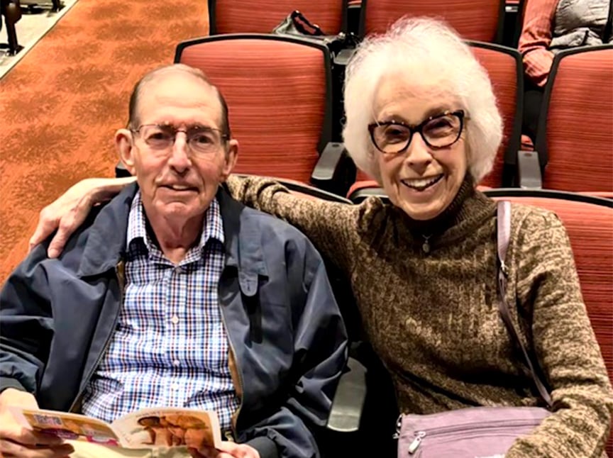 An older man and woman sit together in red theater seats, smiling at the camera. The woman has her arm around the man. He holds a program and wears glasses, a plaid shirt, and a jacket; she wears glasses and a brown sweater.