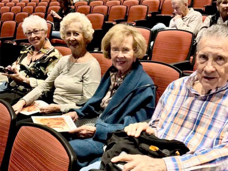 Four older adults sit together in a theater with red seats, smiling at the camera. Two women hold programs or booklets. Other people are seated in the background.