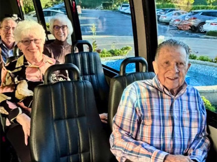 Four older adults are sitting and smiling inside a small bus with black seats. The bus windows show a parking lot and greenery outside.