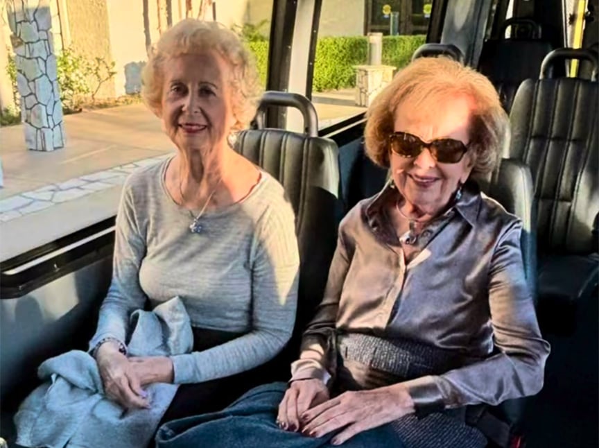 Two elderly women sit side by side on a bus. One woman wears a light sweater and holds a blanket, while the other wears sunglasses and a shiny blouse. They both smile, enjoying the sunlight streaming through the windows.
