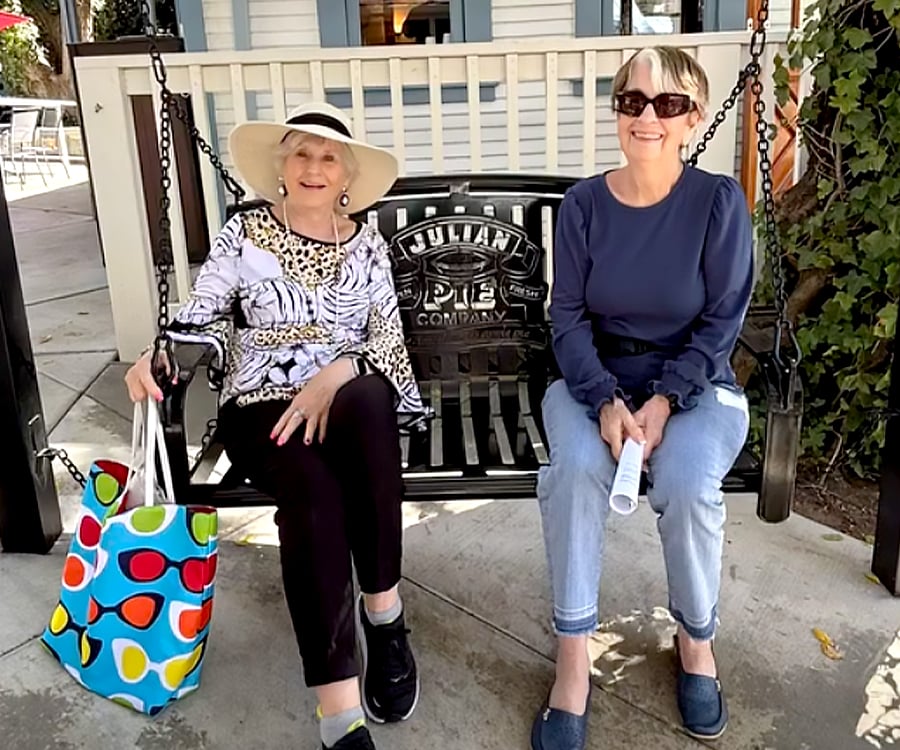 NNC-COV_Hero_Dec2025 Two women sit smiling on a black metal swing bench. One wears a wide-brimmed hat and holds a colorful tote bag, while the other wears sunglasses and holds a rolled-up paper. They are outdoors on a sunny day.