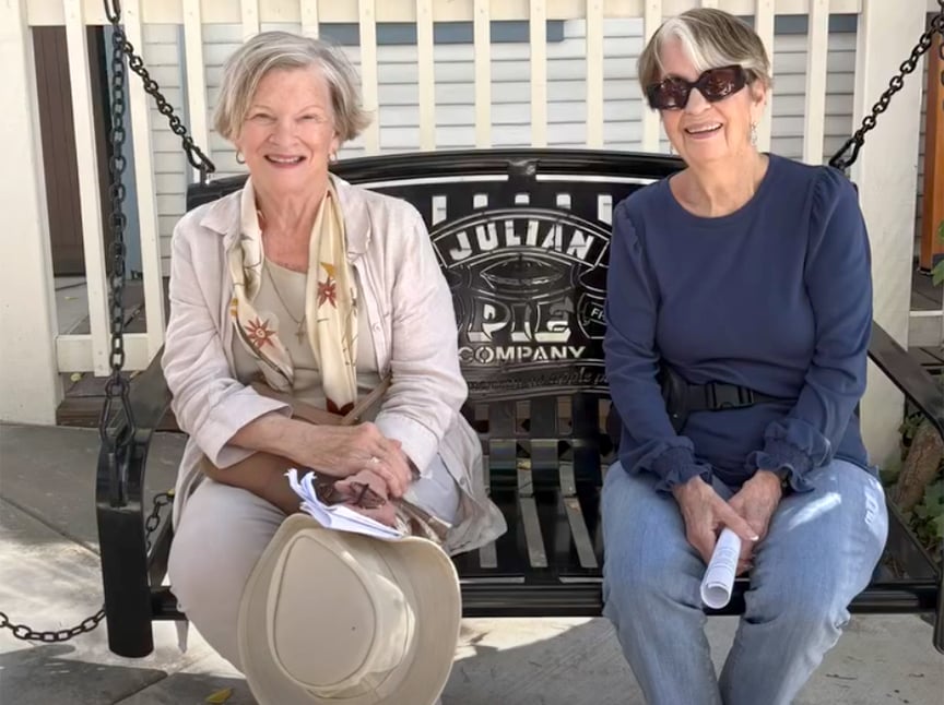 Screenshot Two older women sit and smile on a black metal bench with Julian Pie Company on the backrest. One holds a hat and papers; the other holds a rolled-up paper. Both wear casual outfits and sunglasses. A white fence is behind them.