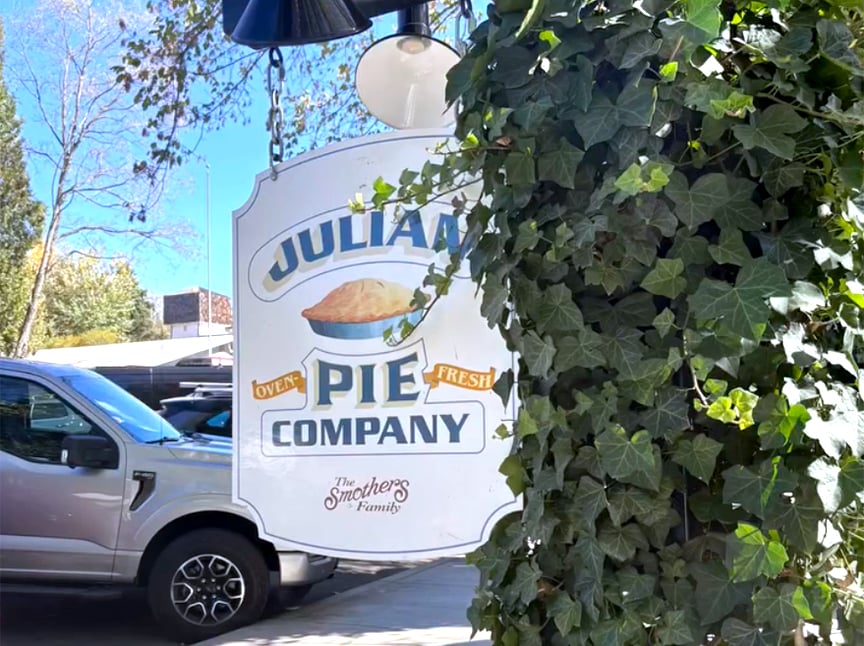 Screenshot A sign for Julian Pie Company hangs outside, partially covered by ivy. The sign features an image of a pie and the words Oven Fresh. A silver pickup truck is parked nearby under a clear, sunny sky.
