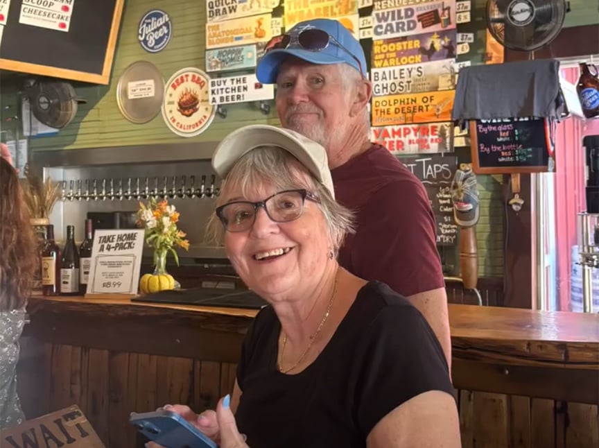 Screenshot Two older adults, a woman in glasses and a white cap and a man in a blue cap, smile at the camera while standing at a bar with colorful beer signs and taps in the background.