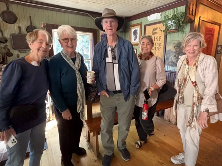 Screenshot Five older adults, four women and one man, stand indoors smiling at the camera. They are casually dressed; the man wears a hat. The cozy room features wooden floors, vintage decor, and framed pictures on the walls.