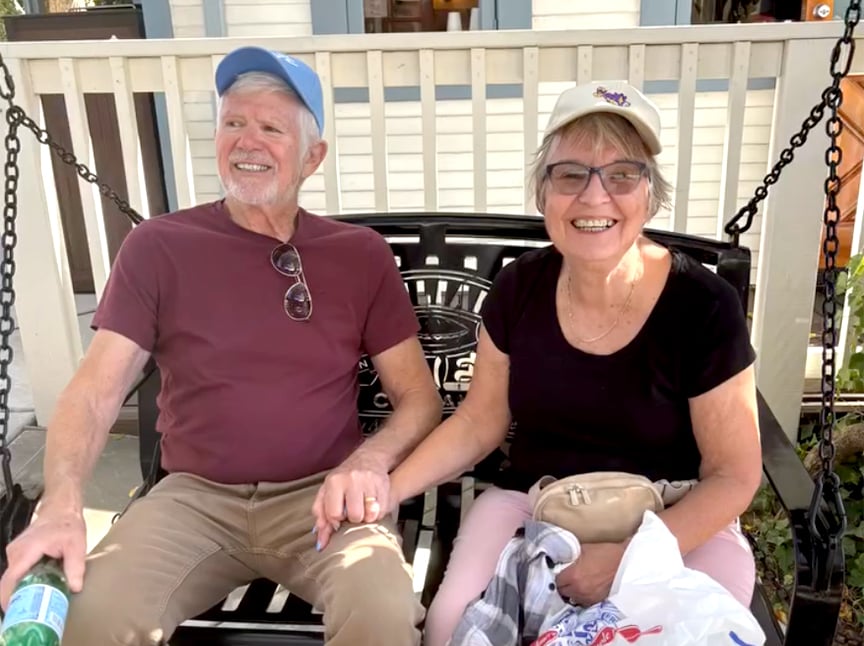 Screenshot An older man and woman sit together on a bench swing outside, smiling and holding hands. Both wear glasses and baseball caps. The woman holds a bag and a jacket. They look happy and relaxed.