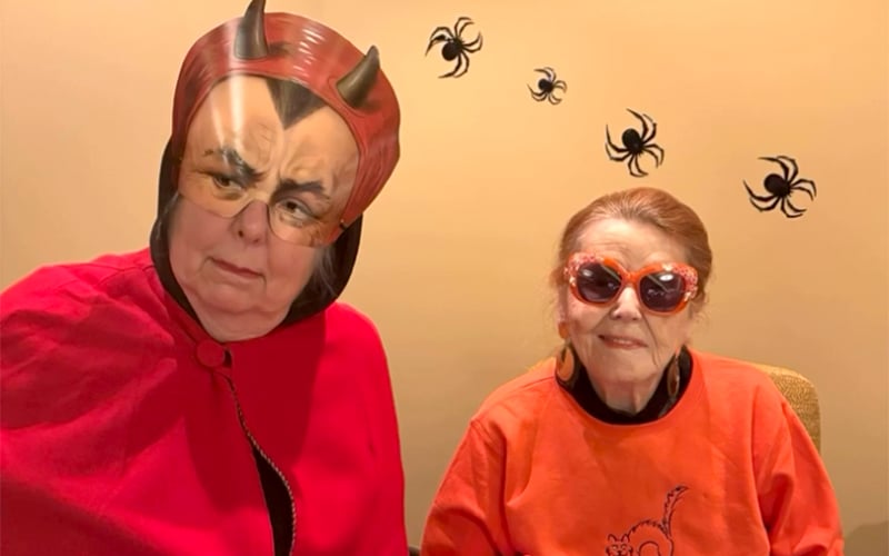 Screenshot Two older women in Halloween costumes: one wears a devil mask with horns and a red cape, the other has heart-shaped sunglasses and an orange shirt. Black spider decorations are on the tan wall behind them.
