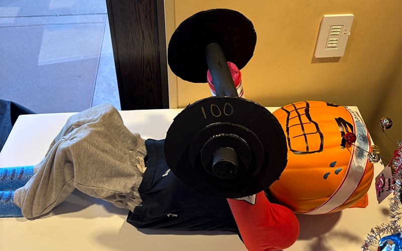 Screenshot A pumpkin decorated like a person lies on a table, dressed in clothes and socks, lifting a barbell labeled 100 in a playful scene. The pumpkin has a face drawn on it and wears a helmet.