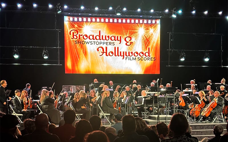 Screenshot An orchestra performs on stage in front of an audience, with a large illuminated screen behind them reading Broadway Showstoppers & Hollywood Film Scores.
