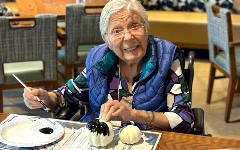 Screenshot An elderly woman with gray hair, glasses, and a colorful shirt smiles while painting a small white pumpkin black. She sits at a table with art supplies, holding a brush and a pumpkin, in a brightly lit room.