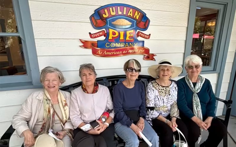 Screenshot Five older women sit smiling on a bench in front of the Julian Pie Company sign, which features a pie and the slogan As American as apple pie. They are wearing casual clothes and hats.