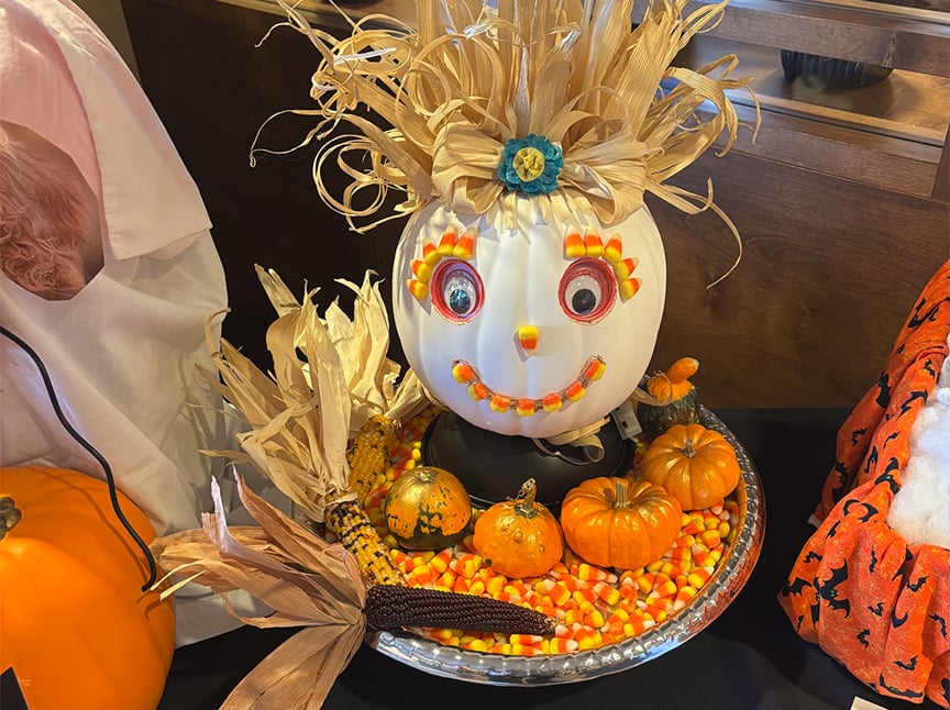 Screenshot A white pumpkin decorated with corn husks for hair, candy corn for facial features, and button eyes sits on a platter surrounded by mini pumpkins, decorative corn, and candy corn pieces.