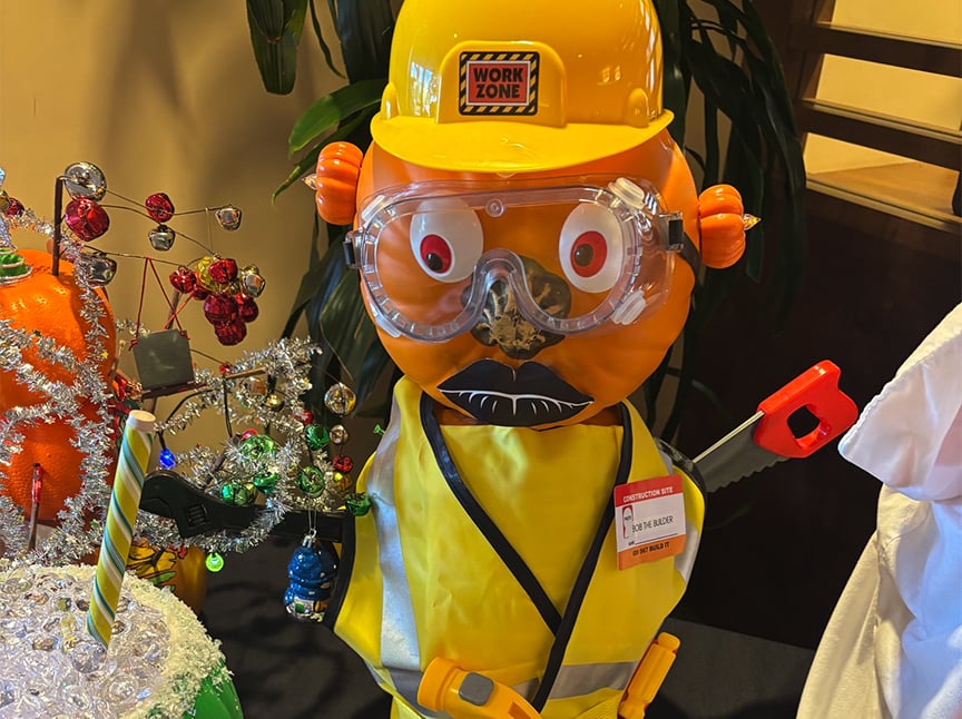 Screenshot A pumpkin decorated as a construction worker, wearing a yellow hard hat, safety goggles, safety vest, and ID badge. The pumpkin has cartoonish eyes and mouth, and holds a toy saw.