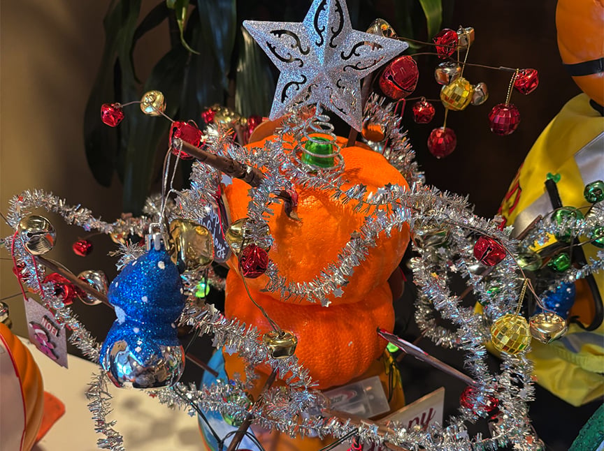 Screenshot A small artificial Christmas tree decorated with tinsel, colorful ornaments, and a silver star topper stands in front of an orange pumpkin. The scene features bright, festive holiday decorations.