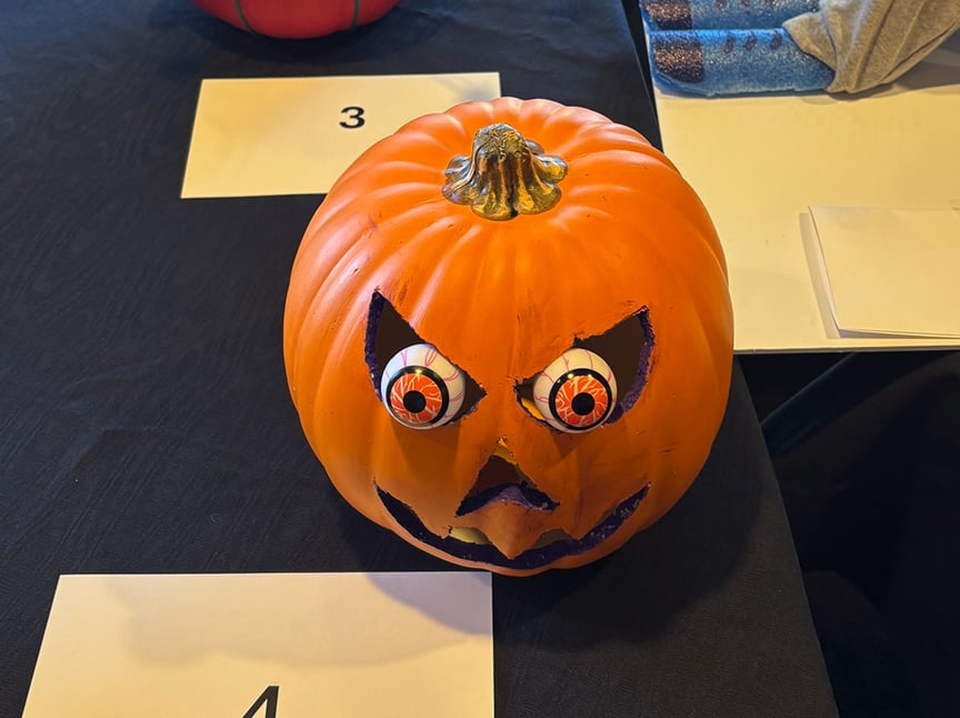 Screenshot A carved pumpkin with exaggerated, bulging red eyes and a jagged mouth sits on a black tablecloth. A white card with the number 4 is placed in front of it.