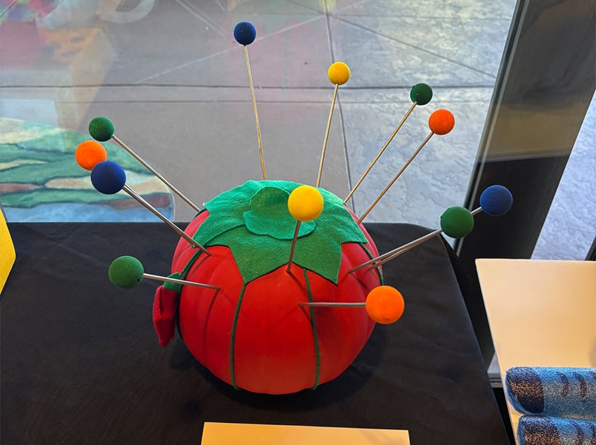 Screenshot A large red tomato-shaped pincushion topped with green felt leaves, holding long pins with colorful ball heads in blue, yellow, orange, and green, sits on a black surface near a window.