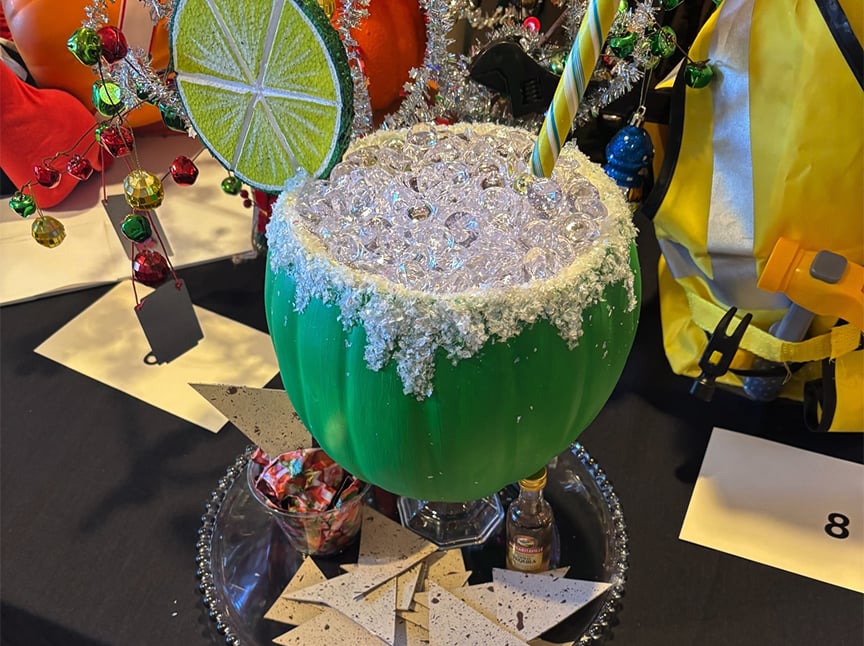 Screenshot A large green decorative cup with faux ice, a lime slice, and a striped straw sits on a tray, surrounded by festive holiday ornaments, small bottles, and candy.