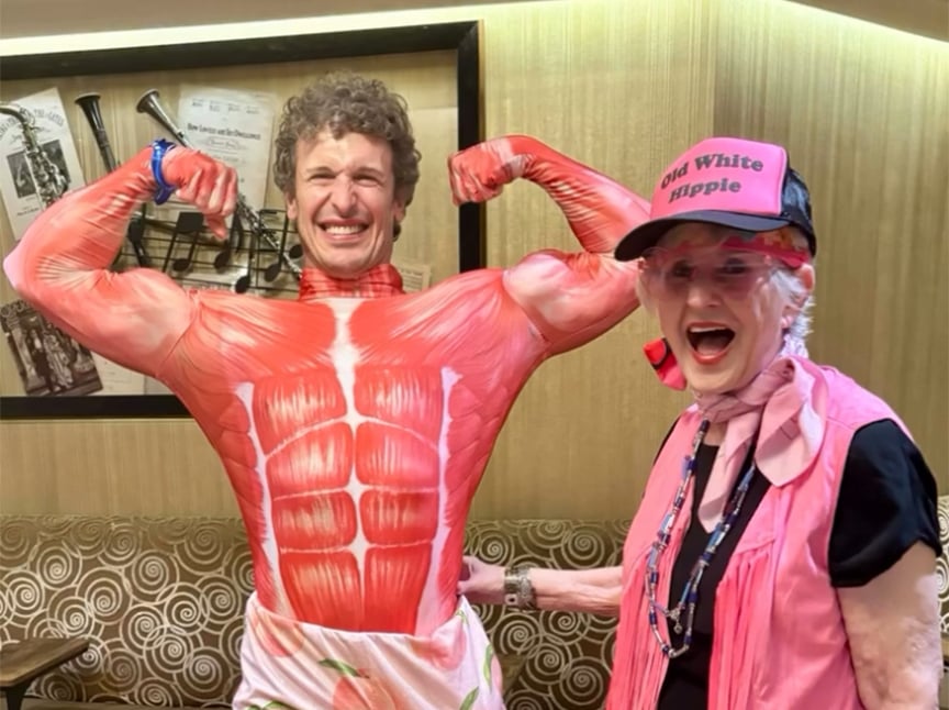 Screenshot A man wears a muscle suit and flexes his arms, smiling widely, while an older woman in a pink cap that reads Old White Hippie, pink scarf, and sunglasses stands beside him, also smiling. They appear to be having fun indoors.