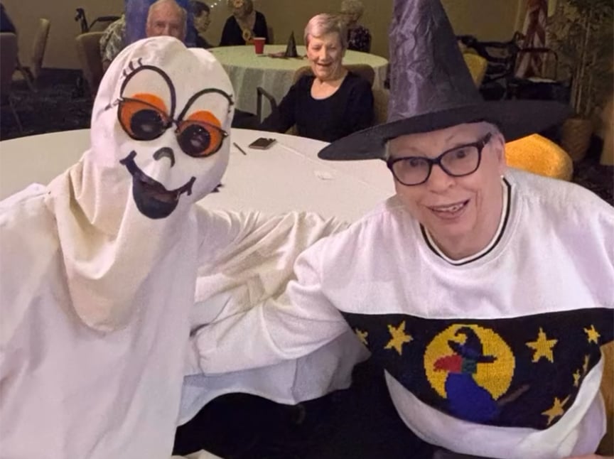 Screenshot Two people in Halloween costumes pose together at a table. One wears a ghost outfit with sunglasses, and the other wears a witch hat and a sweater with a witch design. Several other people sit at tables in the background.