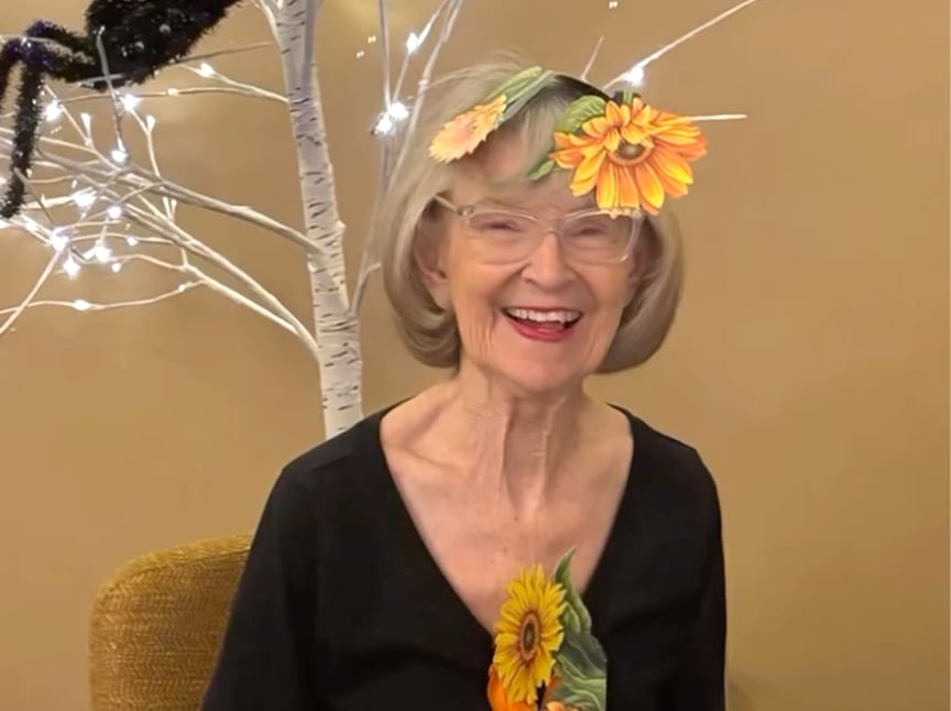 Screenshot Smiling older woman with glasses sits in front of a white tree with lights and a large fake spider. She wears a black top and has virtual sunflowers on her head and chest, likely from a filter.