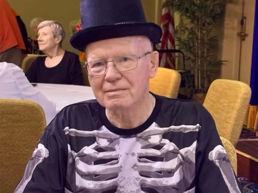 Screenshot An older man wearing glasses, a black top hat, and a shirt with a skeleton print sits at a table. Another older woman sits in the background. The setting appears to be a decorated indoor gathering.