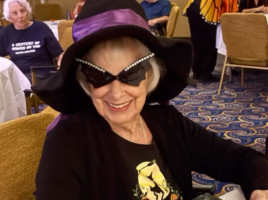 Screenshot An elderly woman wearing a large black hat with a purple ribbon and jeweled cat-eye sunglasses smiles while seated indoors on patterned carpet. People are visible in the background.